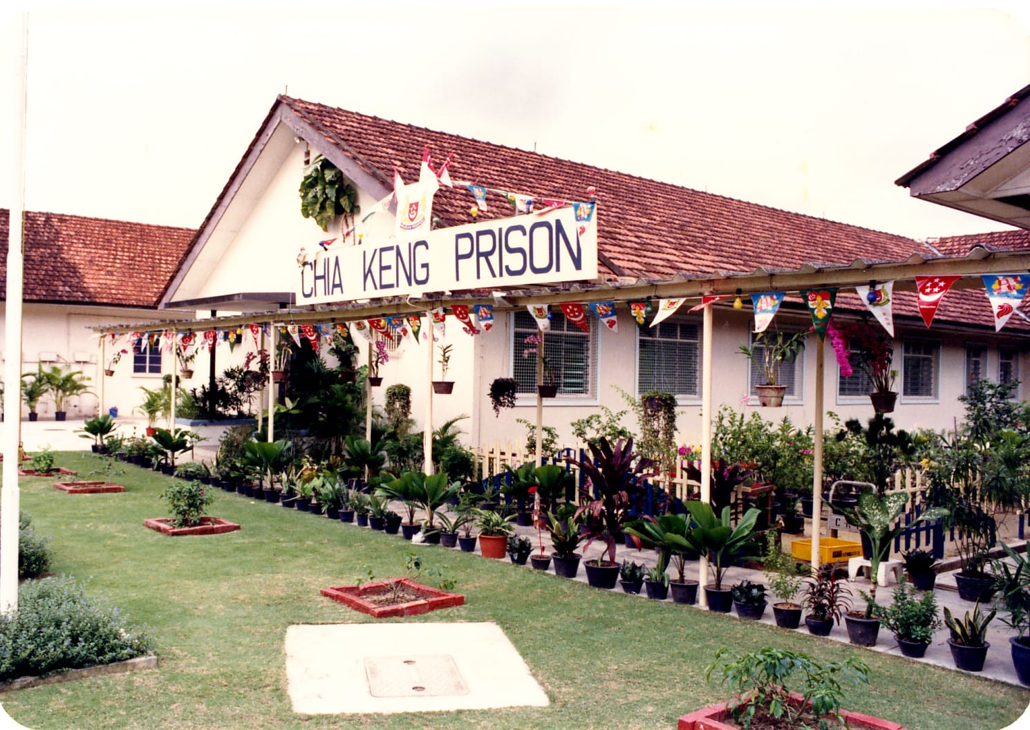 A beige building with a sign reading "CHIA KENG PRISON," adorned with colorful flags and surrounded by potted plants.